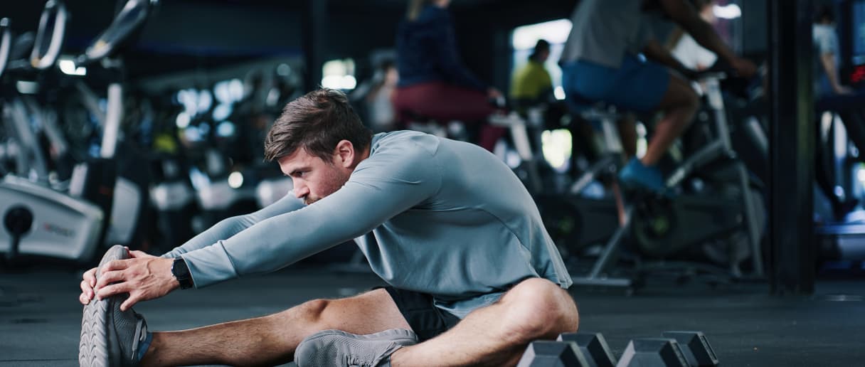 Sportista isteže nogu sedeći u modernoj teretani, pored kardio sprava i tegova.